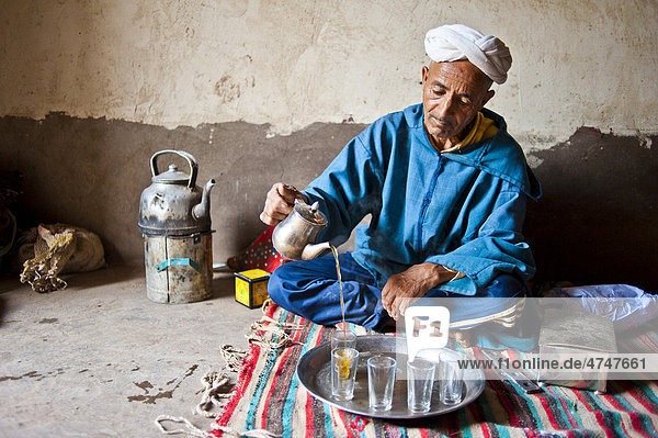 Ein älterer Berber mit Turban hockt am Boden auf einer Decke und schenkt aus einer Silberkanne den traditionellen Minzetee aus Kelaa M'gouna Hoher Atlas Marokko Afrika