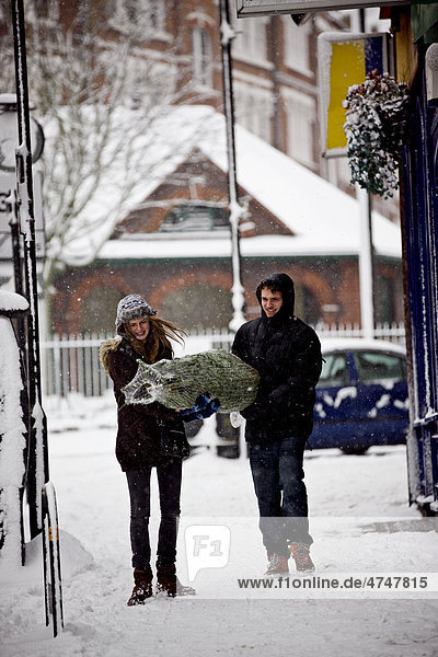 Ein junges Paar trägt einen Weihnachtsbaum auf einer verschneiten Straße