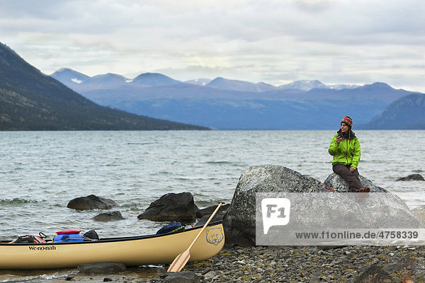 Junge Frau sitzt auf einem Felsen  am Ufer liegt ein Kanu mit Holzpaddel  am See Kusawa Lake  dahinter die Berge  Yukon Territorium  Kanada