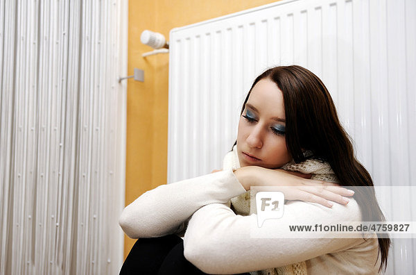 Young woman sitting and shivering in front of a heater  heating costs