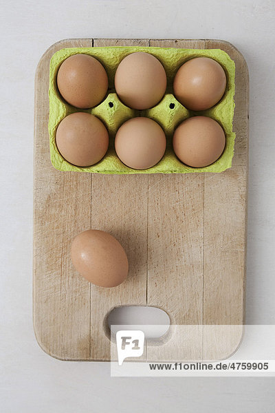 Six brown eggs in an egg carton and one egg on a wooden cutting board beside it