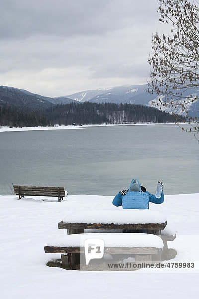 Young woman sitting at a picnic table  searching for something inside her bag on the lakeshore of Walchensee or Lake Walchen  picnic table  winter  Bavaria  Germany  Europe