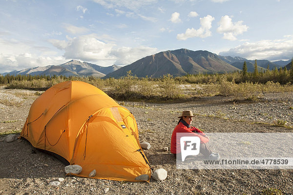 Junge Frau sitzt entspannt vor ihrem Zelt und genießt die Abendsonne  dahinter die Northern Mackenzie Mountains  Wind River  Yukon Territory  Kanada