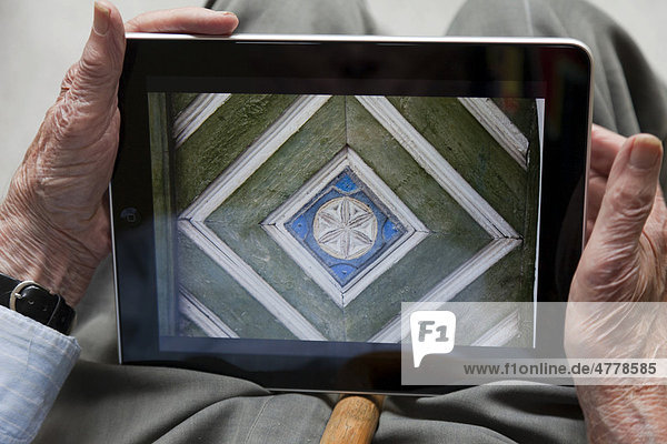 Senior betrachtet Foto einer alten Holzschnitzerei aus dem Unterengadin  Schweiz  auf seinem iPad  Nahaufnahme Hände