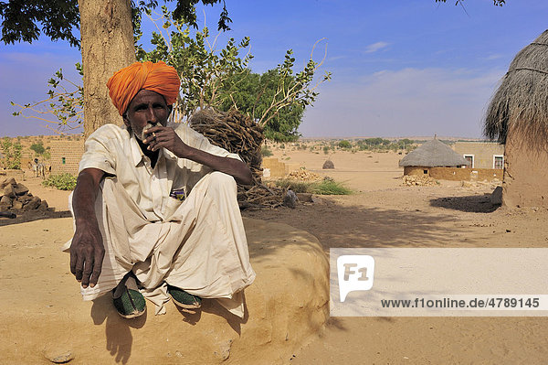 Inder mit Dohti  traditionelles Beinkleid  und Turban hockt rauchend auf einer Lehmbank neben seinem Haus  Wüste Thar  Rajasthan  Indien  Asien