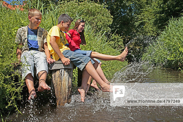 Young people sitting on a dock beside a stream and splashing water with their bare legs  Bad Bodenteich  Lower Saxony  Germany  Europe