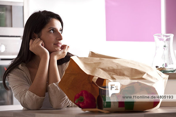 Young woman sitting at a dining table in the kitchen  thinking  paper bag containing purchases at front