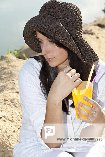 Young woman wearing a straw hat sitting on the beach of a lake and holding a refreshing drink in her hand