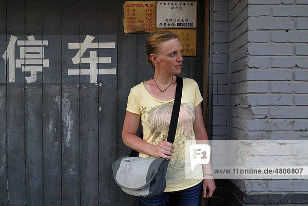 Red-haired woman carrying a shoulder bag standing in front of a house on a Chinese Hutong  a narrow street  Chinese characters behind her  Beijing  China  Asia