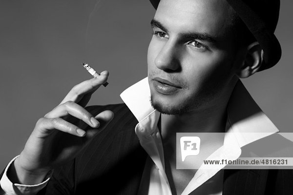 Junger Mann mit Anzug  Hemd  Krawatte und Hut hält eine Zigarette in seiner Hand