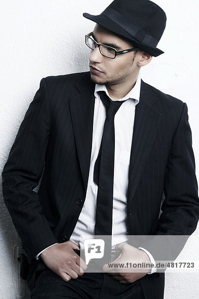 Portrait of a young man wearing glasses  a suit  a shirt  a tie and a hat leaning against a wall