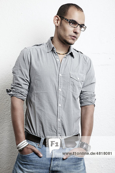 Young man wearing glasses  jeans and a shirt  leaning against a wall with his hands in his pockets