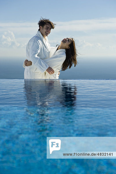 Couple embracing and laughing at edge of infinity pool  both wearing bathrobes