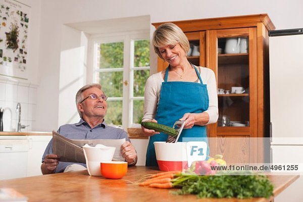 Älterer Mann mit Zeitung  ältere Frau bei der Zubereitung des Essens