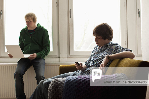 A teenage boy on a laptop while his friend text messages on his mobile