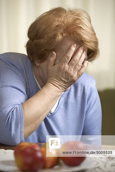 Eine ältere Frau sitzt an einem Tisch und bedeckt ihr Gesicht mit ihrer Hand.
