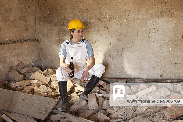 Eine Bauarbeiterin sitzt auf Trümmern und hält ein Bier.