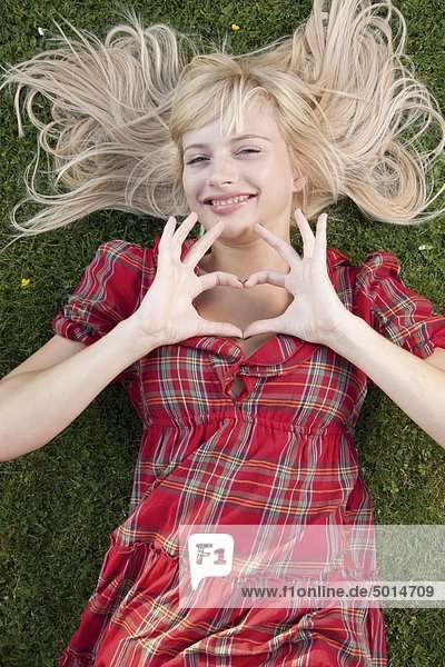 Lächelnde blonde Frau im Gras formt ein Herz mit ihren Händen
