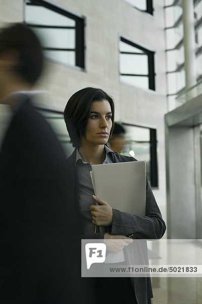 Professional woman standing in lobby  looking away in thought as executives walk past