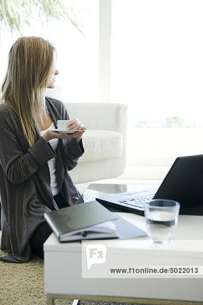 Frau entspannt im Wohnzimmer mit Tasse Kaffee und Laptop  Blick aus dem Fenster