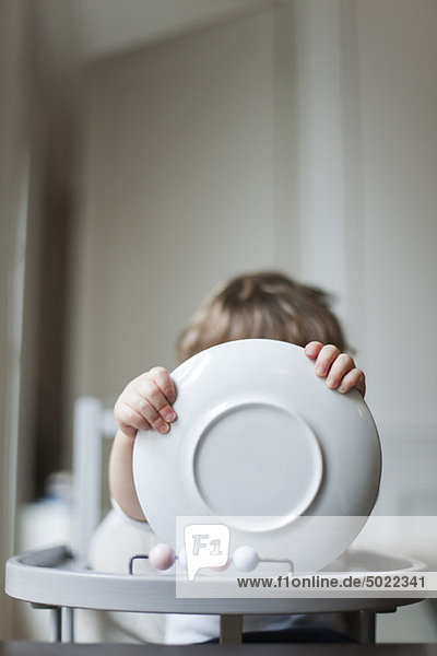 Toddler sitting in high chair  holding plate in front of face