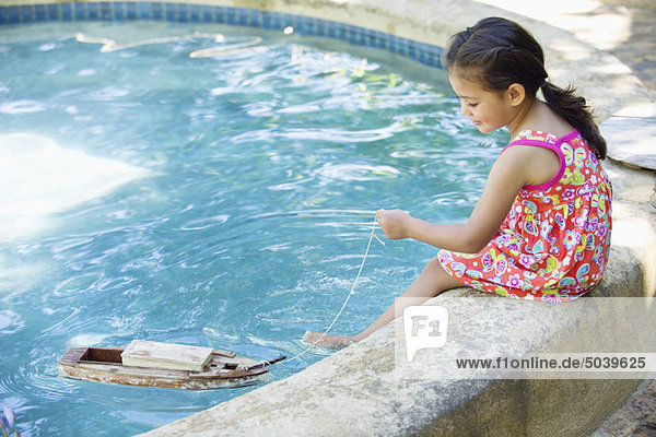 Mädchen sitzt am Rand des Schwimmbades und schaut auf ein Spielzeugboot im Wasser.