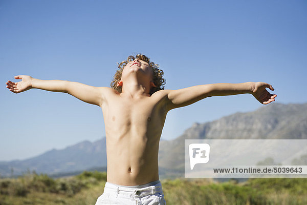 Ein kleiner Junge ohne Hemd  der mit geschlossenen Augen und ausgestreckten Armen gegen den Berg steht.