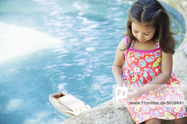 Mädchen sitzt am Rand des Schwimmbades und schaut auf ein Spielzeugboot im Wasser.