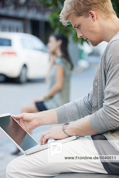 Young man using a laptop with a woman in the background on a street