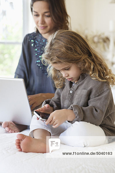 Mädchen sieht sich ein Smartphone an  während ihre Mutter an einem Laptop auf dem Bett arbeitet.