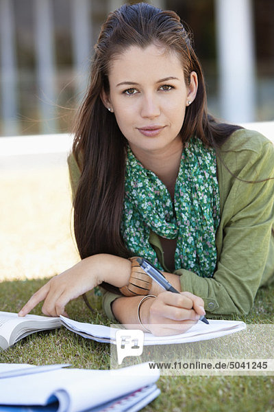 Porträt einer jungen Frau  die auf Gras liegt und auf einem Campus studiert.