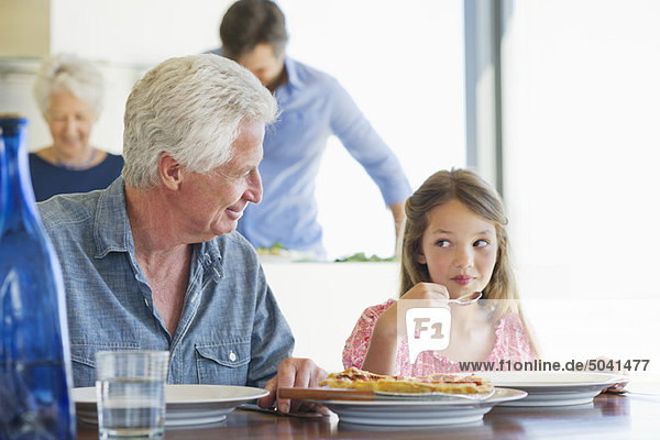 Girl eating food at a dining table with her grandfather sitting near her