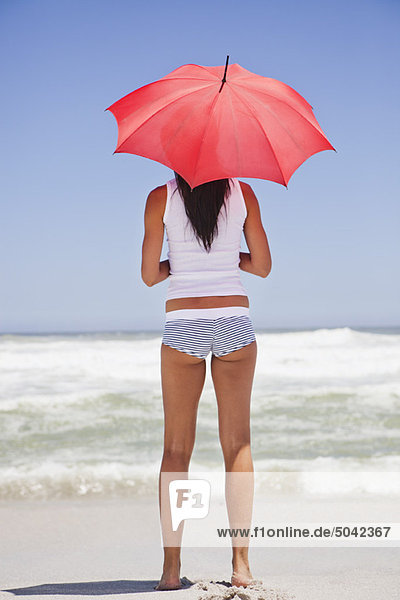 Frau am Strand stehend mit Sonnenschirm und Blick aufs Meer
