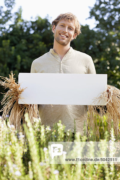 Young man in scarecrow dressing and holding a blank placard in a field