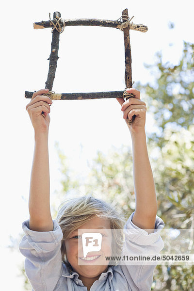 Portrait of a cute little boy holding frame of driftwood with arms raised outdoors