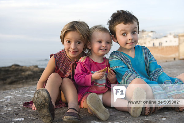 Junge Geschwister sitzen zusammen am felsigen Strand  Porträt