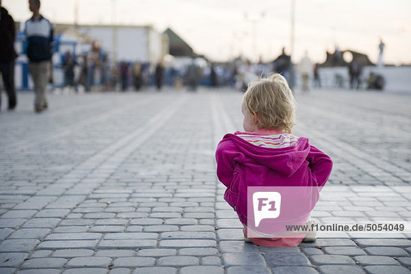 Kleinkind-Mädchen sitzt auf dem Boden auf dem Stadtplatz und sieht zu  wie Leute vorbeigehen.