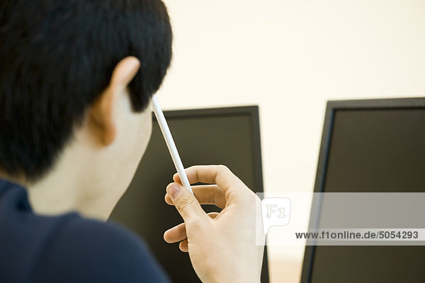 Junger Mann schaut auf Computerbildschirme  hält Bleistift gegen Kopf in Gedanken