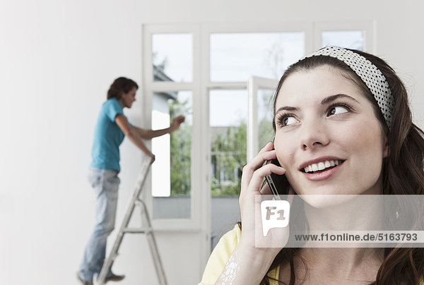 Nahaufnahme einer jungen Frau mit Handy und Männermalerei im Hintergrund bei der Wohnungsrenovierung