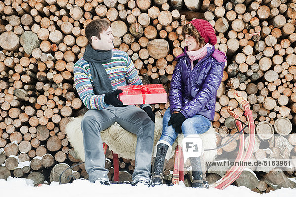 Österreich  Salzburger Land  Flachau  Junger Mann verschenkt Weihnachtsgeschenk an Frau auf Schlitten mit Brennholz im Hintergrund
