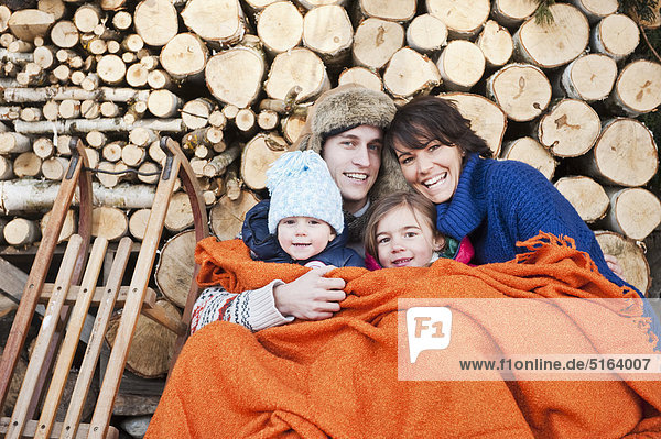 Österreich  Salzburger Land  Flachau  Familie unter Wolldecke sitzend vor Feuerholz