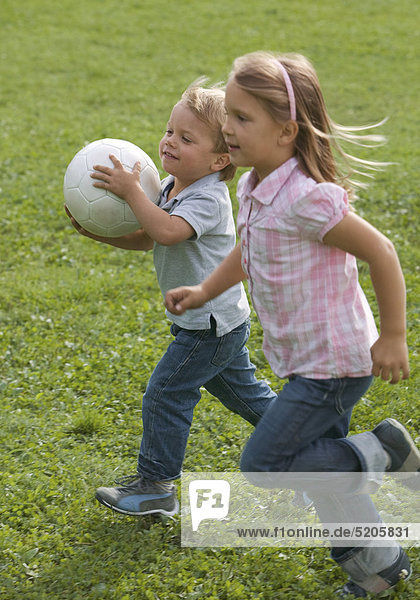 Kleiner Junge und Mädchen rennen mit Fußball über Wiese