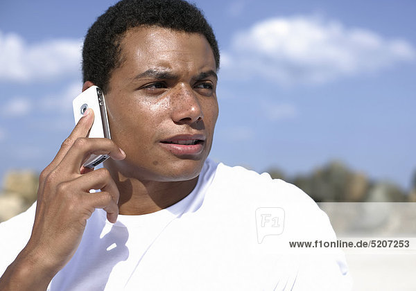 Mann  dunkelhäutig  weißes T-Shirt  telefoniert mit Handy am Strand