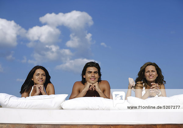 Ein Mann und zwei Frauen liegen an Strand auf Kissen unter freiem Himmel