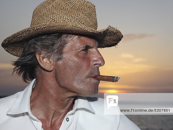 Älterer Mann mit Strohhut und Zigarre vor Sonnenuntergang am Meer