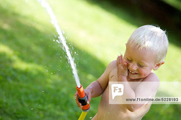 Kleiner blonder Junge spritzt mit Wasser im Garten