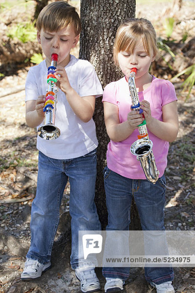A boy and a girl standing by a tree and playing toy musical instruments