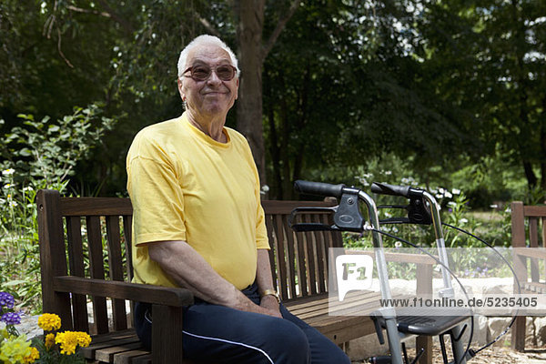 Ein älterer Mann mit einem Geher  der auf einer Bank im Hof seines Pflegeheims ruht.