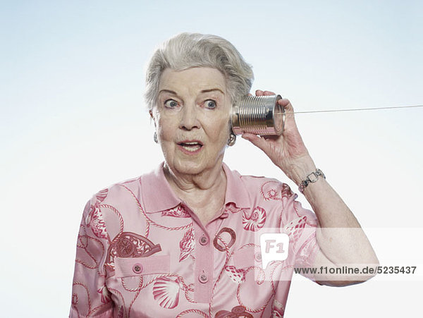 Seniorenfrau hört komisch zu  als sie eine Blechdose an den Kopf hält.