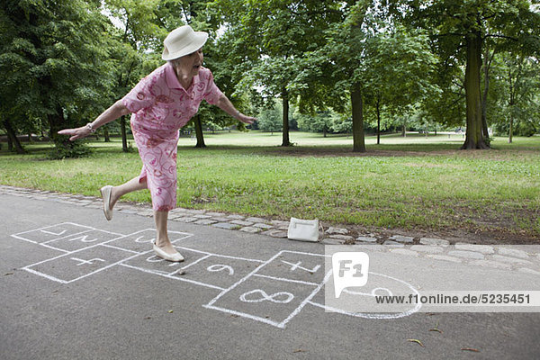 Seniorin spielt Hopscotch und versucht  ihr Gleichgewicht zu halten.